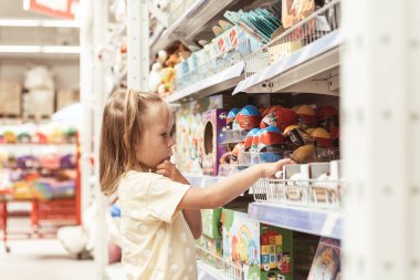 Küçük beyaz kız büyük bebek mağazasında yeni bir oyuncak seçiyor. Oyuncak dolu büyük raflar