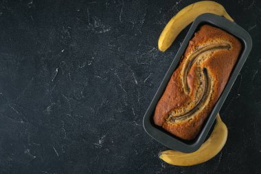 Siyah bir yemek kabında taze muzların yanında siyah beton bir arka planda taze muzlu turta. Ev yapımı hamur işi konsepti. Düz yatıyordu. Boşluğu kopyala