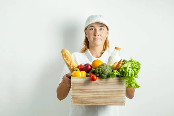 Young caucasian delivery woman in white uniform with grocery wooden box with fresh fruits and vegetables isolated on white background. Shopping and delivery service concept.