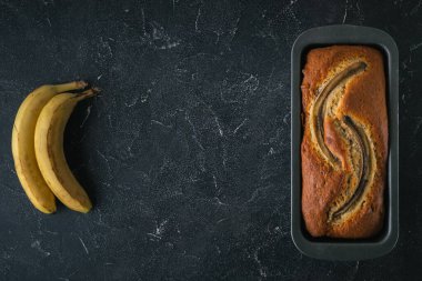 Siyah bir yemek kabında taze muzların yanında siyah beton bir arka planda taze muzlu turta. Ev yapımı hamur işi konsepti. Düz yatıyordu. Boşluğu kopyala