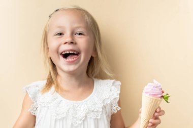 Beautiful little caucasian girl with blonde hair in a white dress enjoying berry flavored waffle cone ice cream on the beige background