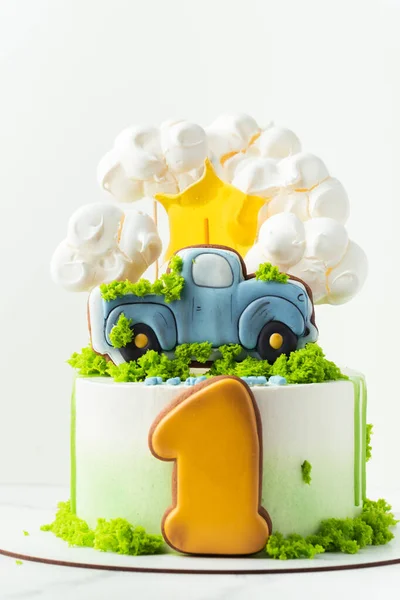 Birthday cake for a one year old kid. White cake decorated with green edible grass and gingerbread cookies in the shape of a truck and digit one. Fluffy cloud shaped meringue cookies on top of cake