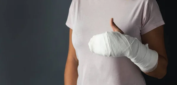 Woman with a cast on her arm showing thumbs up on the dark grey background. Girl has a broken hand wearing pink T-shirt