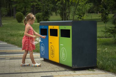 Şirin küçük kız parktaki sarı konteynıra plastik çöp atıyor. Çöp ayıklama seti. Cam, kağıt ve plastik çöp için sembol dolu kutular. Atık ayıklama yoluyla çevrenin bakımı