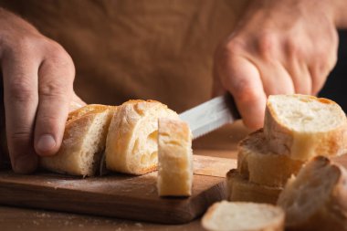 Whole grain bread close-up on the wooden cutting board. Man cutting fresh bread with a big knife. Knife with serrated blade. Fresh bread on the kitchen table. Traditional pastries in bakery