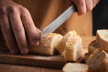 Whole grain bread close-up on the wooden cutting board. Man cutting fresh bread with a big knife. Knife with serrated blade. Fresh bread on the kitchen table. Traditional pastries in bakery
