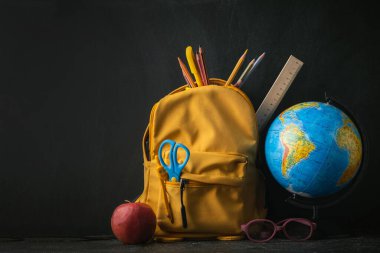Yellow backpack with school supplies next to the globe, red apple and glasses on the black school board background. Back to school concept on September day