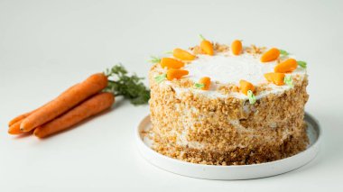 Delicious carrot cake decorated with mastic sweet carrots. Homemade carrot cake with yellow crumbs in the white plate on the white background