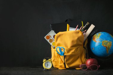 Yellow backpack with school supplies next to the globe, red apple and glasses on the black school board background. Back to school concept on September day