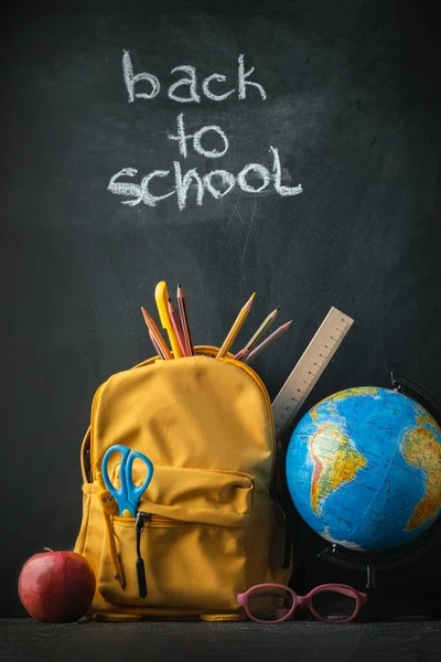 Yellow backpack with school supplies next to the globe, red apple and glasses on the black school board background. Back to school concept on September day