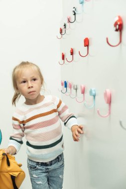 Cute little caucasian girl hanging yellow backpack on a white hanger in the school locker room. Back to school concept