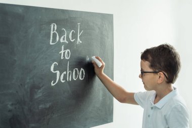 Cute caucasian schoolboy wearing glasses and writing Back to school text on the black school board in the classroom at elementary school.