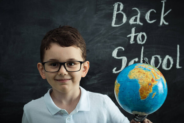 Cute caucasian schoolboy wearing glasses and holding the Globe against the black school board in the classroom at elementary school. Back to school concept