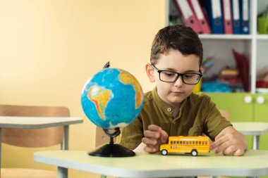 Cute caucasian schoolboy wearing glasses and sits at a desk in the classroom at elementary school. Little boy next the Globe playing with yellow school bus toy. Back to school concept
