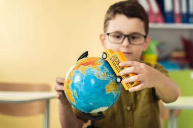 Cute caucasian schoolboy wearing glasses and sits at a desk in the classroom at elementary school. Little boy next the Globe playing with yellow school bus toy. Back to school concept