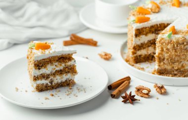 Delicious carrot cake decorated with mastic sweet carrots next to white cup of tea. Slice of homemade carrot cake with yellow crumbs in the white plate on the white background