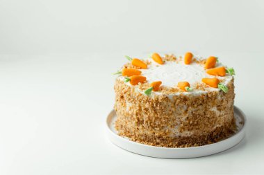 Delicious carrot cake decorated with mastic sweet carrots. Homemade carrot cake with yellow crumbs in the white plate on the white background