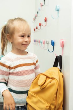 Cute little caucasian girl hanging yellow backpack on a white hanger in the school locker room. Back to school concept