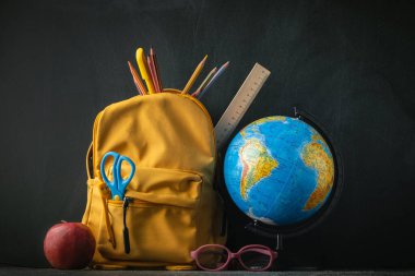 Yellow backpack with school supplies next to the globe, red apple and glasses on the black school board background. Back to school concept on September day