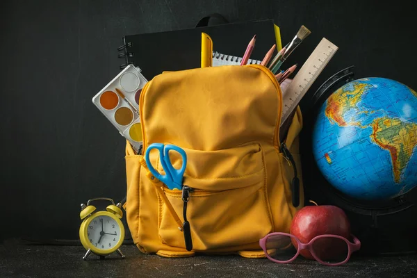 Yellow backpack with school supplies next to the globe, red apple and glasses on the black school board background. Back to school concept on September day