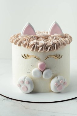 Doğum günü pastası beyaz krem peynirli krem şantili mastik kedi kulakları, pençeler ve yüzle süslenmiş. Beyaz arka plandaki küçük bir kız için sürpriz pasta.