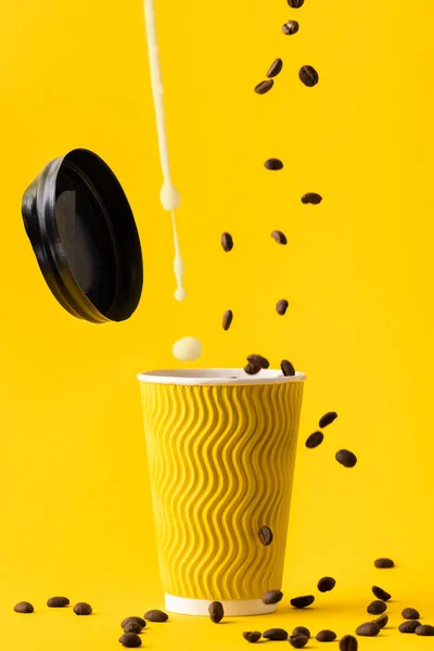 Coffee beans and milk drops falling in the yellow paper cup on the yellow background. Making cup of coffee in the morning. Levitation of coffee drink ingredients