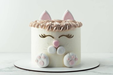 Doğum günü pastası beyaz krem peynirli krem şantili mastik kedi kulakları, pençeler ve yüzle süslenmiş. Beyaz arka plandaki küçük bir kız için sürpriz pasta.