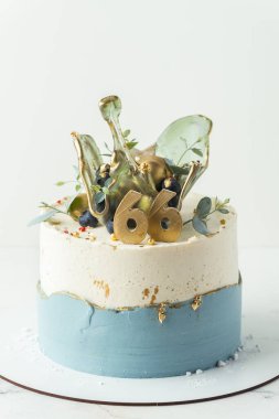 Beyaz ve mavi krem peynirli bir yıl dönümü pastası üzerinde şeffaf karamel sıçrama figürü ve 66 rakamlı altın çikolata.
