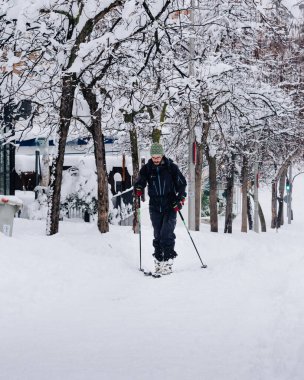 Yoğun kar yağarken ve devrilmiş ağaçlarla kaplı bir caddede kayak yapan bir adam.