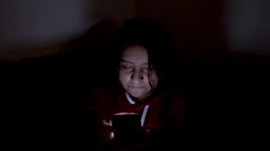 Close up shot of a girl sitting on a couch in dark and checking her phone. Mobile screen light is flashing on her face.