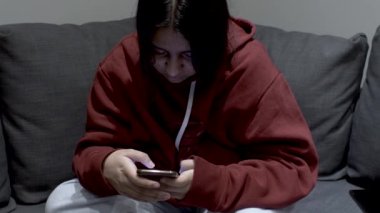 A teenage youth dressed in a hoody sitting on a sofa at home, the young girl focused on her mobile phone as she messages with a group of friends
