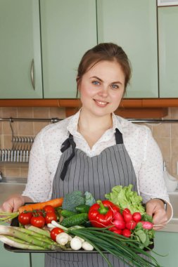 Mutlu Kadın mutfak arka planında taze sebze malzemeleriyle dolu bir tepsi tutuyor. Sağlıklı vejetaryen yemekleri. 