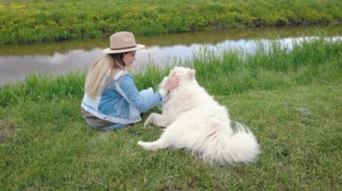 Genç ve güzel bir kadın oturmuş beyaz köpeğini kucaklıyor nehrin yakınındaki bir doğa tarlasında.