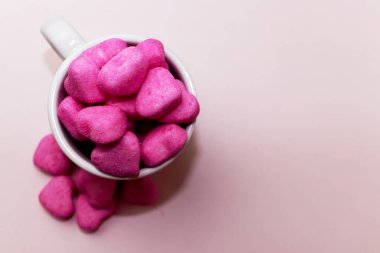 Pembe arka planda beyaz bir fincanda pembe kalp şeklinde şekerler yığını.