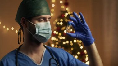 Koronavirüs aşısıyla maskeli erkek doktor. Arka planda noel ışıkları var. 