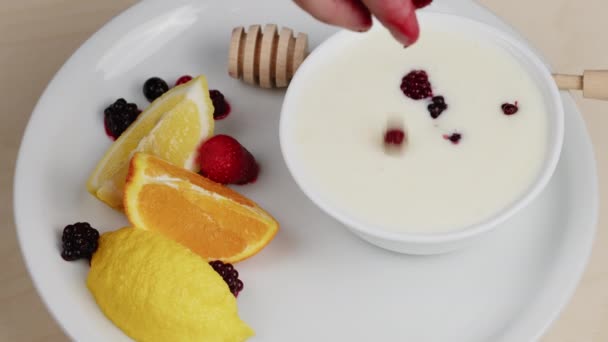 Mão coloca mirtilos de frutas vermelhas em uma xícara cheia de iogurte branco — Vídeo de Stock
