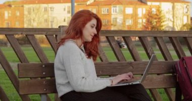 Dizüstü bilgisayarda çalışan genç bir kız öğrenci üniversitede ahşap bir bankta oturuyor.