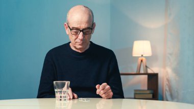 Gözlüklü yaşlı adam evdeki masada oturmuş kalp hapı yiyor.