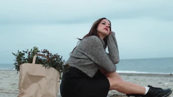 Chica joven sentada en la playa cerca del mar con un montón de hojas — Vídeo de stock