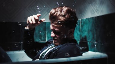 Mutlu iş adamı takım elbise ve kravatla küvette sabun köpüğüyle oynar. El kamerasıyla çekim.