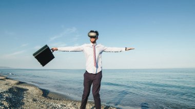 Business guy spins on himself with virtual reality glasses on the beach near the sea. Static shoot.