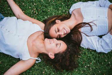 İki genç kız parkta çimlerin üzerinde uzanırken gülümseyip birbirlerine bakıyorlar. Rahatlamış görünüyorlar..