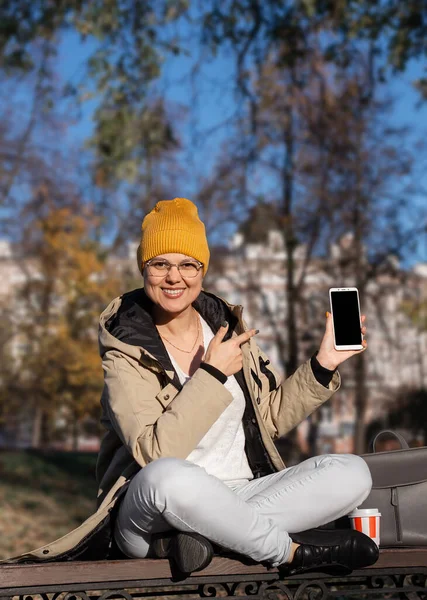 Młoda uśmiechnięta kobieta siedzi ze skrzyżowanymi nogami i wskazuje na ekran smartfona w dłoni. Skupienie selektywne — Zdjęcie stockowe