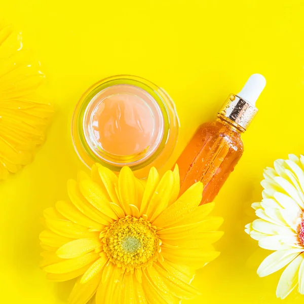 Vitamin C serum and open pink cream with yellow beautiful flowers on monochrome background in water. Skin care products flat lay, square