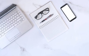 Düz dizüstü bilgisayar, akıllı telefon ve gözlüklü beyaz mermer masa üstünde çeşitli kırtasiyeler.