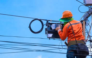 Ahşap merdivendeki teknisyenlerin arka görüntüsü internet dağıtıcı kutusundaki fiber optik kablo hatlarının kod numaralarını kontrol ederek mavi gökyüzü arka planına karşı elektrik direğinde normal çalışması için onarım yapmak.