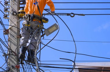 Güvenlik ekipmanları ve çeşitli iş araçlarına sahip elektrikçilerin bir kısmı mavi gökyüzü arkaplanına karşı elektrik direğine kablo hattı kurmak için çalışıyor 