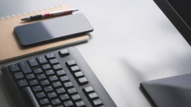 Akıllı telefonun yüzeyinde sabah ışığı, tükenmez kalem, bilgisayar klavyesinin bir parçası olan spiral defter ve masa başı çalışma alanındaki monitör