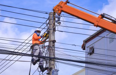 Vinçli koruyucu maskeli Asyalı elektrikçi mavi gökyüzü arka planına elektrik kabloları ve elektrik iletimi kuruyor.