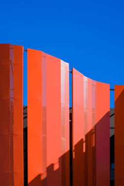 Yüksek binanın dışındaki turuncu kıvrımlı çelik duvar dekorasyonunun yüzeyindeki güneş ışığı ve gölge dikey çerçevede mavi gökyüzüne karşı.